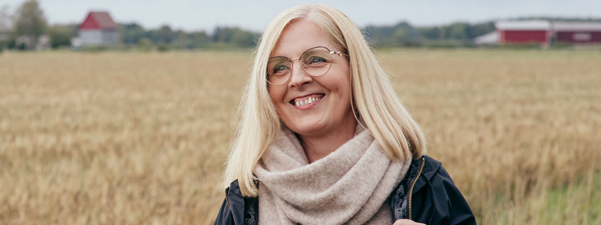 Rouva hymyilee pellon laidalla, kasvoilla kultaiset silm&auml;lasit